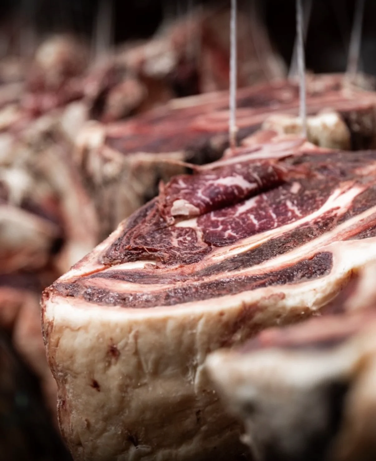Pièce de viande persillée suspendue à des crochets métalliques, avec d’autres morceaux en arrière-plan flou.